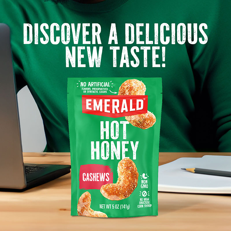 Green pack of Emerald Hot Honey Cashews in front of a person wearing a green sweater, next to a laptop and notebook. Text above reads, Discover a Delicious New Taste! Package highlights non-GMO and no artificial ingredients.